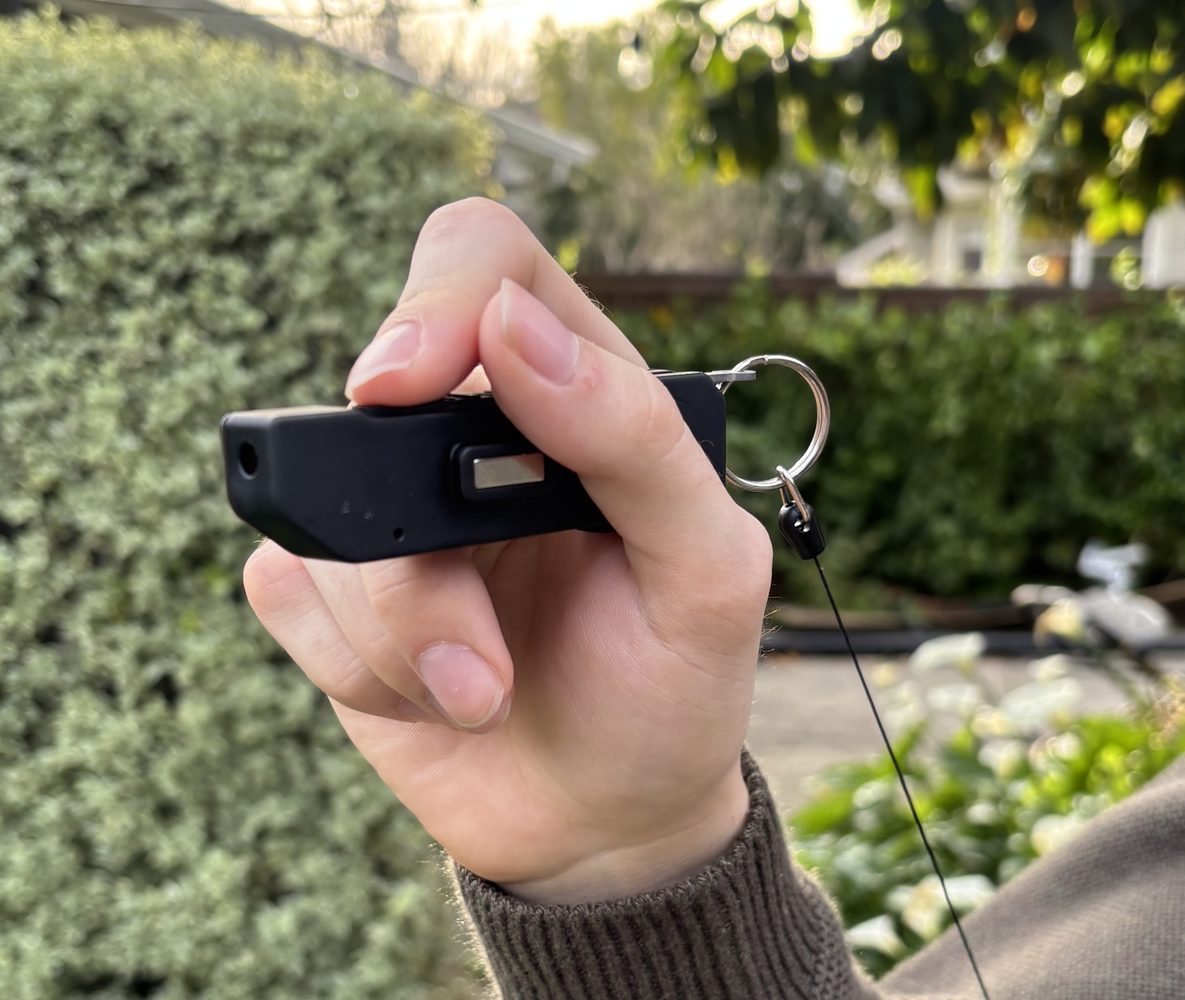 A hand holding the black Glance wearable outdoors, with a thumb resting near its controls.